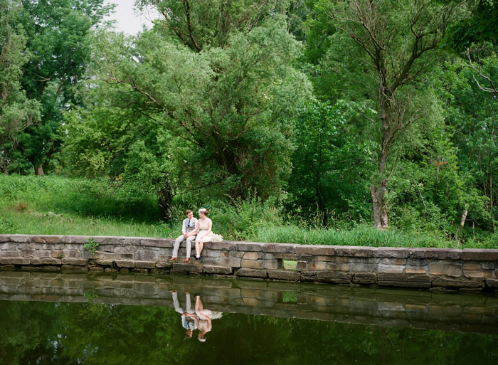 bride and groom sitting at edge of pond