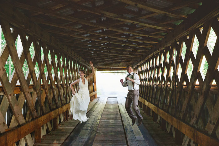 bride and groom jumping on covered bridge