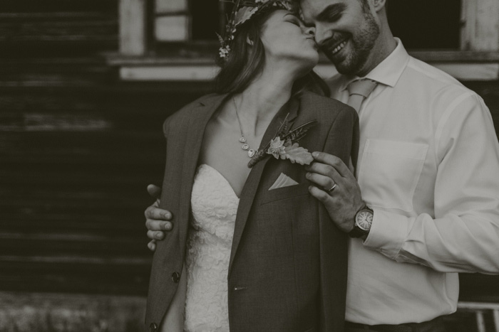 bride wearing groom's jacket and kissing him on the cheek