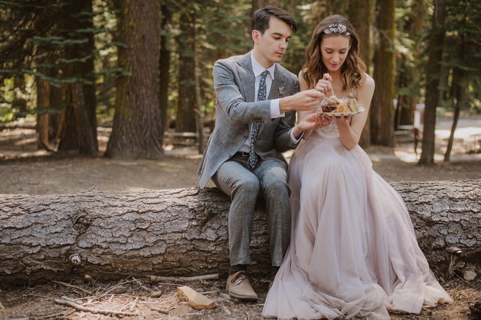 bride and groom eating cake on a log in the woods